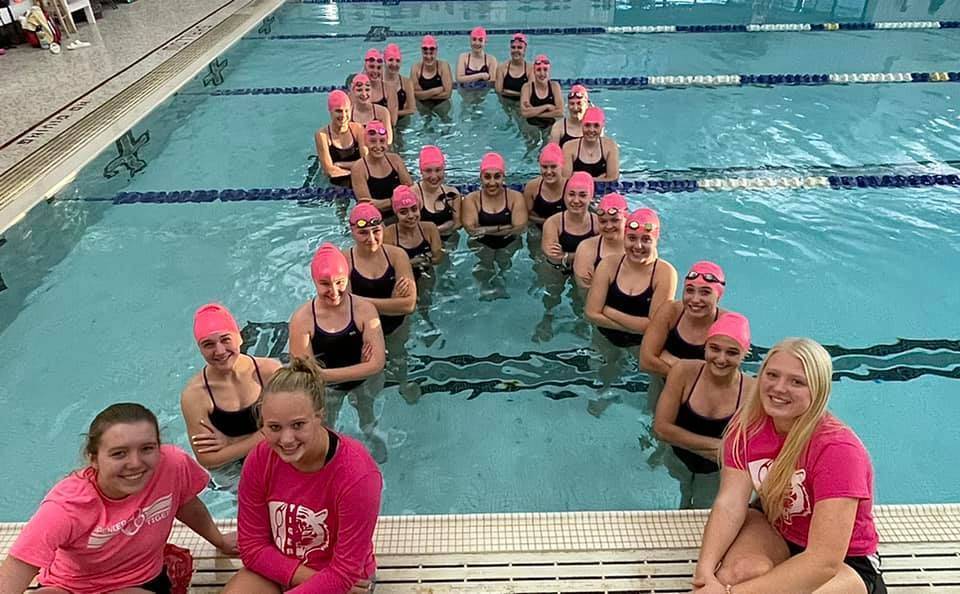 Swimmers wearing pink swim caps form a ribbon shape in the pool